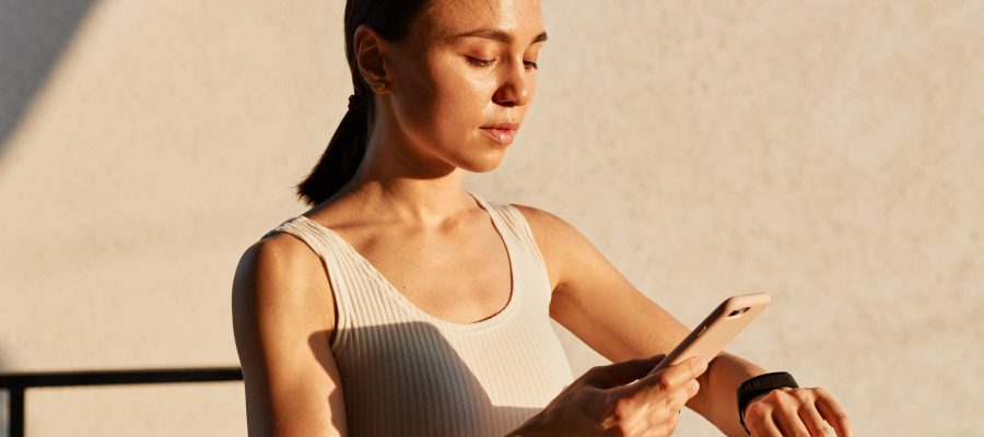 Beautiful brunette female with ponytail wearing white top, connecting her smart phone with fitness bend on her wrist to monitor burned calories after working out.